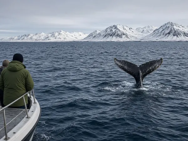 A humpback whale tail rising from the sea beside a small boat, with snowy mountains behind
