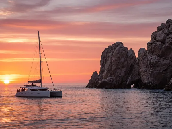 A white catamaran sailing past the Land's End rocks at sunset under an orange and pink sky